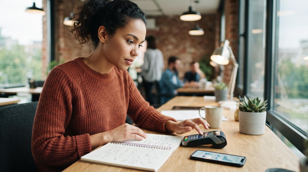 Uma pessoa analisando anotações em um caderno aberto enquanto usa uma maquininha de cartão ao lado de um smartphone, com expressão concentrada